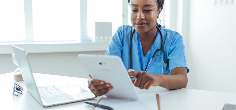 COLOUR-IMAGE_black-female-nurse-with-braids-using-white-tablet-sat-at-desk_apr26