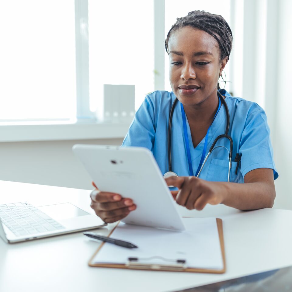 COLOUR-IMAGE_black-female-nurse-with-braids-using-white-tablet-sat-at-desk_apr26