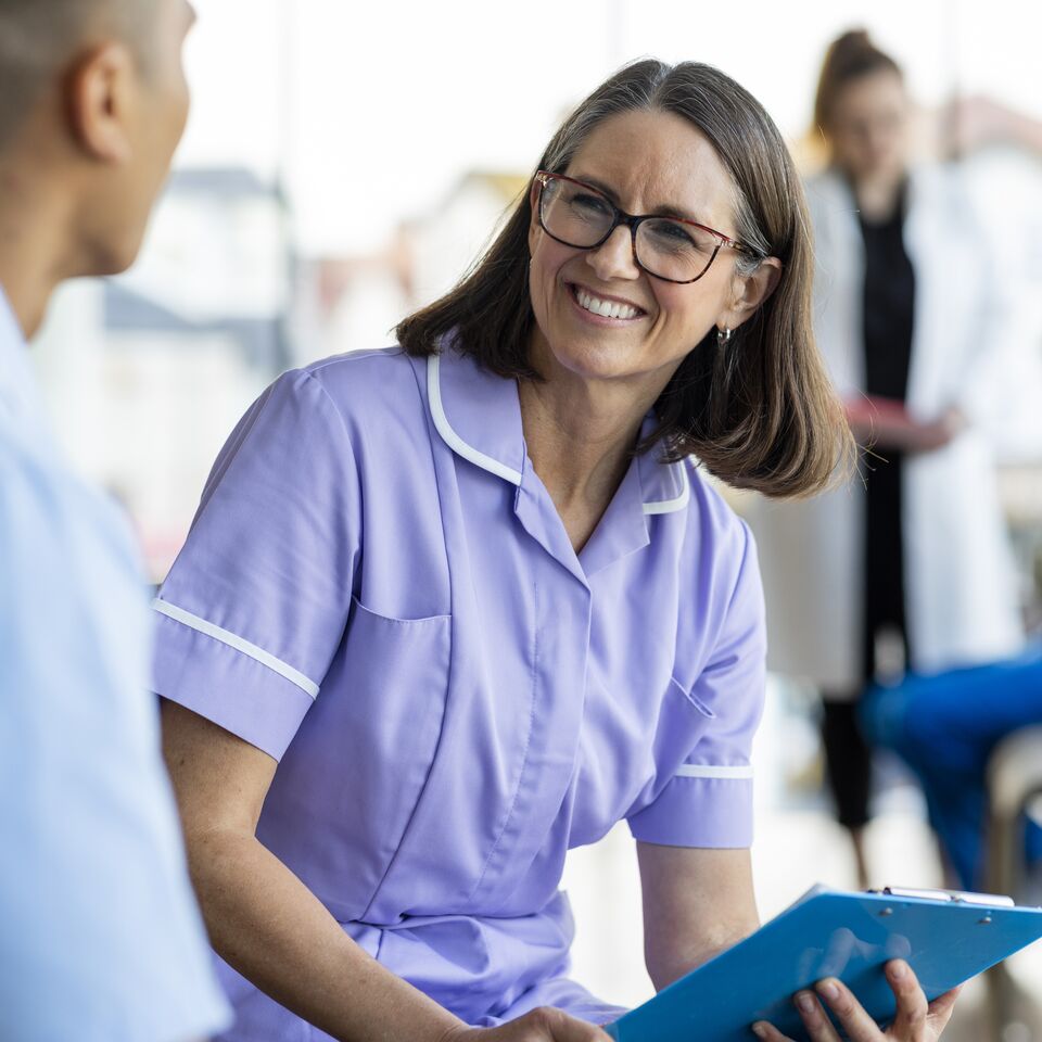 COLOUR-IMAGE_older-female-nurse-in-lilac-scrubs-smiling-at-male-nurse_apr26