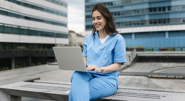COLOUR-IMAGE_brunette-female-nurse-using-laptop-on-outdoor-building-terrace_apr26