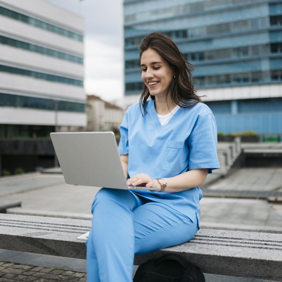 COLOUR-IMAGE_brunette-female-nurse-using-laptop-on-outdoor-building-terrace_apr26