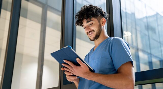COLOUR-IMAGE_dark-haired-male-nurse-stood-against-window-looking-at-tablet_apr26