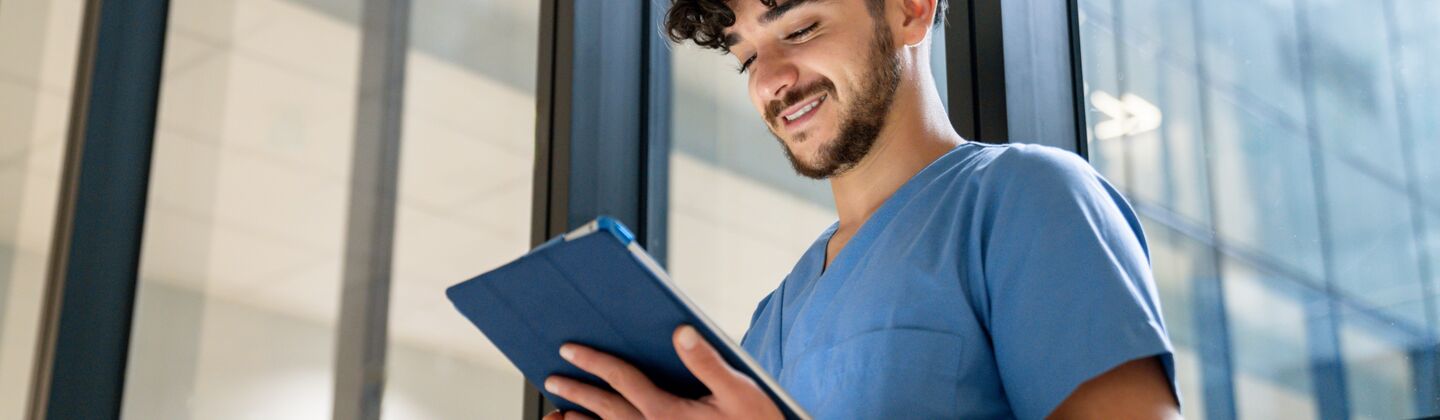 COLOUR-IMAGE_dark-haired-male-nurse-stood-against-window-looking-at-tablet_apr26
