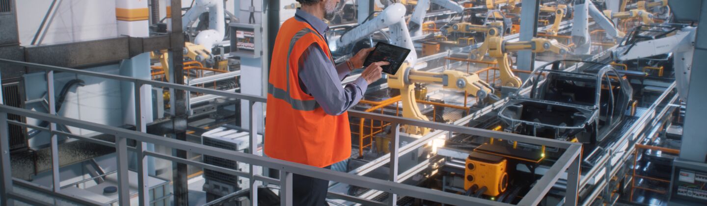 COLOUR-IMAGE_automotive-male-worker-looking-over-automated-robotic-car-assembly-line-holding-tablet_sep26
