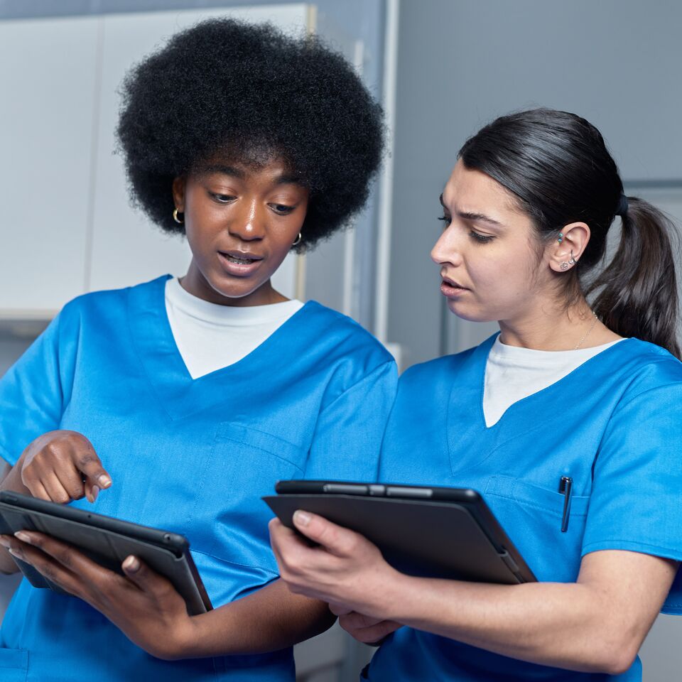 COLOUR-IMAGE_two-female-nurses-in-blue-scrubs-chatting-over-two-tablets_apr26