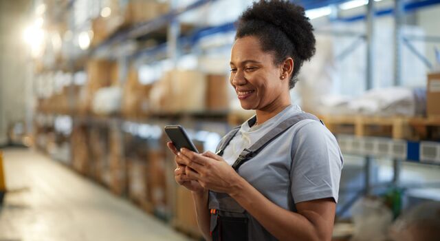 COLOUR-IMAGE_-female-warehouse-worker-smiling-at-mobile-phone_jul26