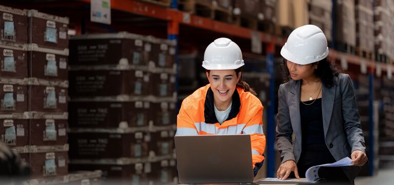 COLOUR-IMAGE-two-female-manufacting-and-supply-chain-workers-with-clipboard-and-laptop-in-warehouse_jul26